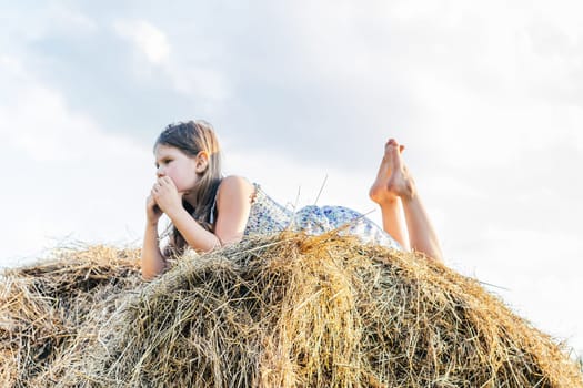 Thoughtful little girl lying on stomach at top of haystack. From below portrait, side view. Child resting on hayrick. Outdoor walking. Beautiful blue sky. Summer vacation, countryside lifestyle.