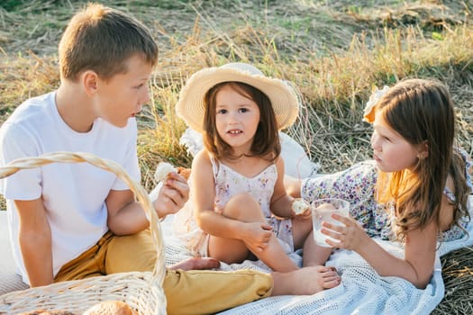 Happy friends talk, eat buns and drink milk sitting on blanket on field. Portrait of little children resting on grass. Outdoor walking and picnic. Summer vacation, countryside lifestyle.