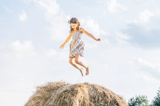 Happy little girl jump up at top of haystack with hands to sides and look down. Joyful kid having fun on hayrick. Outdoor walking. Beautiful blue sky. Freedom. Summer vacation, countryside lifestyle.