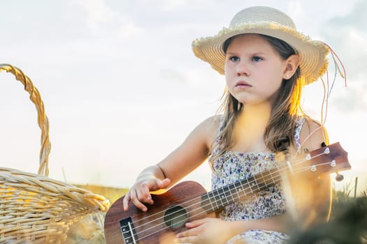 Little girl play ukulele on field. Portrait of child with displeased facial expression in panama. Outdoor picnic. Beautiful sunny sky. Music instrument, hobby. Summer vacation, countryside life.