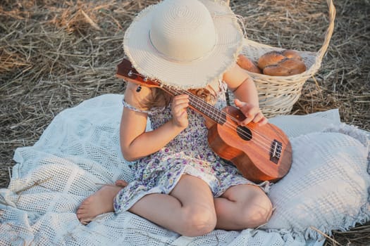 Little girl play ukulele sitting on blanket on field. Portrait of child hiding face under panama hat. Outdoor walking and picnic. Music instrument, hobby. Summer vacation, countryside lifestyle.