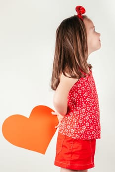Smiling little girl hide red heart shaped greeting card behind back. Portrait of child on white, side view. Give paper cut valentine. Saint Valentine's Day celebration, holiday of all lovers.