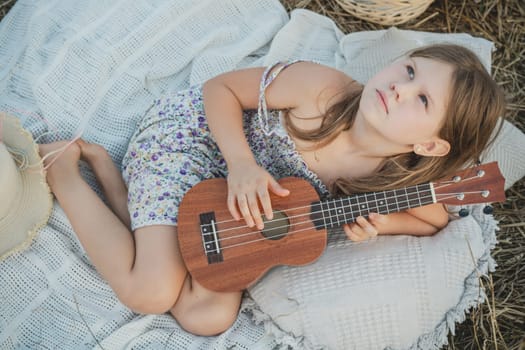 Nice little girl play ukulele lying on blanket on field. Portrait of child looking up. Outdoor walking and picnic. Wooden musical instrument, hobby. Summer vacation, countryside lifestyle.