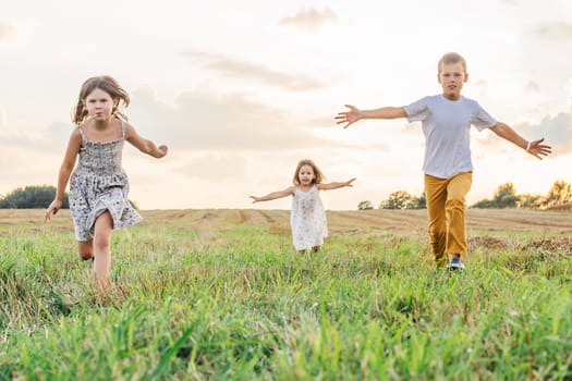 Happy little children joyfully run forward across field waving arms wide. Carefree kids on grassy meadow. Outdoor walking. Beautiful sunny sky. Summer vacation, countryside lifestyle.
