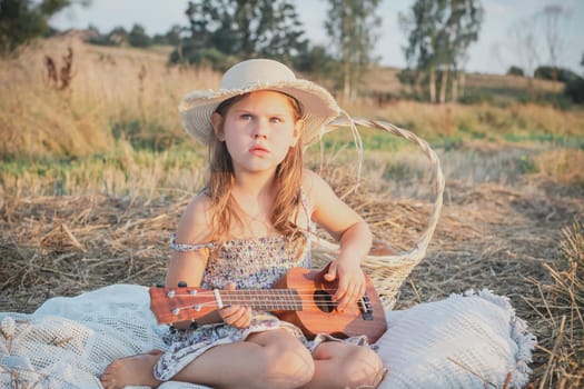 Cute little girl play ukulele sitting on blanket on field. Portrait of child with displeased facial expression in panama. Outdoor picnic. Music instrument, hobby. Summer vacation, countryside life.