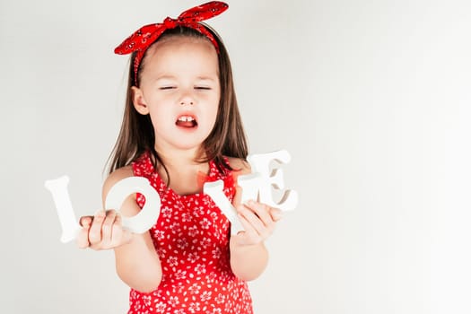 Discontent little girl with closed eyes hold decorative letters LOVE. Portrait of child on white. Kid in festive red outfit. Saint Valentine's Day celebration, holiday of all lovers.