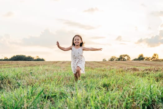 Smiling little girl joyfully running across field. Happy carefree child on grassy meadow. Outdoor walking. Beautiful sunny sky. Summer vacation, countryside lifestyle. Picturesque landscape.