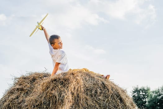 Happy school aged boy sitting at top of haystack launch toy airplane. Carefree child on haymaking play toys. Outdoor walking. Beautiful blue sky. Summer vacation, countryside lifestyle, haying.