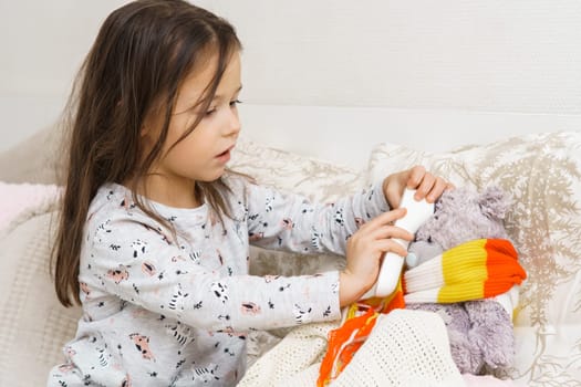 Little girl tie scarf around neck and measure temperature with electronic thermometer of sick teddy bear. Kid in sleepwear sitting on bed and curing ill toy. Fever heat, doctor, treatment.