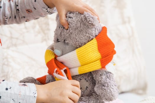 Hands of little girl tie knitted white-orange-yellow scarf around neck of sick teddy bear. Kid in sleepwear playing with stuffed toy. Disease, treatment. Doctor and patient. Closeup photo.