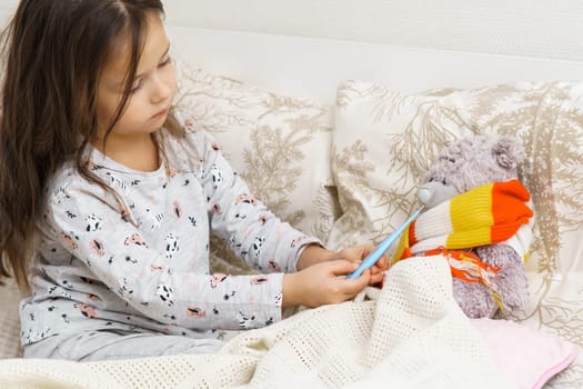 Little girl tie scarf around neck and measure temperature with electronic thermometer of sick teddy bear. Kid in sleepwear sitting on bed and playing with stuffed toy. Fever heat, doctor, treatment.