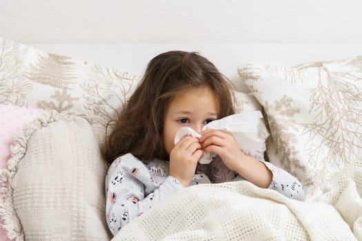 Little girl blow stuffy nose with white handkerchief. Portrait of unhealthy child. Kid in sleepwear lie under blanket and have bed rest. Home treatment. Runny nose, snot, cold symptoms, cure.