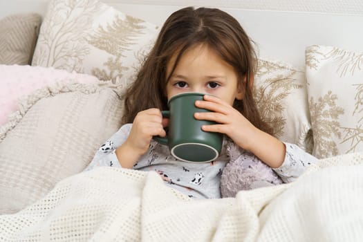 Sick little girl in sleepwear lying under blanket on bed and drink beverage from cup. Portrait of unhealthy child. Kid have bed rest. Home treatment, cold, disease. Taking medicament.
