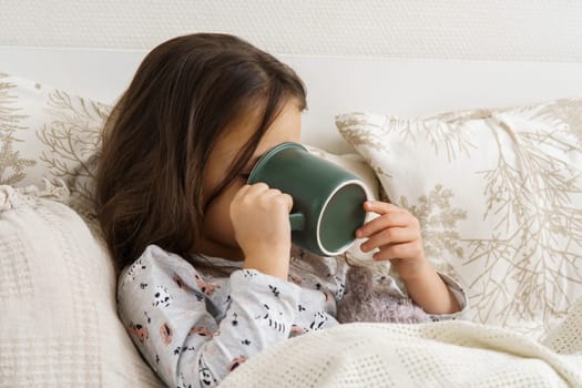 Little girl in sleepwear lie under blanket and take sip of drink covering face with mug. Portrait of unhealthy child, side view. Kid have bed rest. Home treatment, cold, disease. Taking medicament.