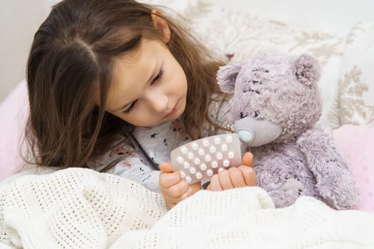 Nice little girl in sleepwear sitting under blanket on couch and treating teddy bear with pills. Portrait of child, preschooler. Kid and toy have bed rest. Home patient treatment. Taking medicament.