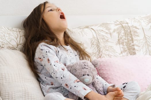 Ill little girl in sleepwear sitting on couch gargle with medicine. Portrait of child, preschooler. Kid and toy have bed rest. Sore throat, home treatment. Taking medicament, cold, disease.