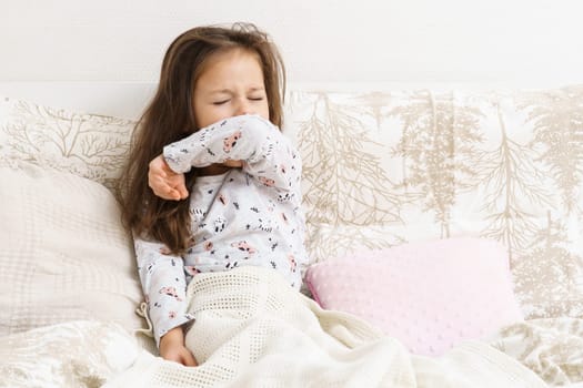 Little girl in sleepwear sit on bed under blanket and yawn with closed eyes covering mouth. Portrait of awaking child. Kid wake up at morning. Cozy bedclothes for sleeping. Resting, awakening.