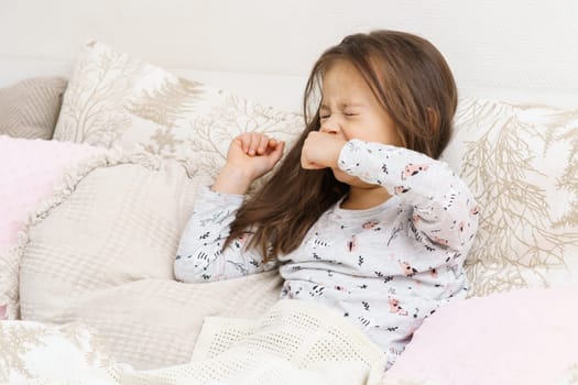 Cute little girl in sleepwear sit on bed under blanket and yawn squinting eyes. Portrait of awaking child. Kid wake up at morning. Cozy bedclothes for sleeping. Resting in bedroom, awakening.