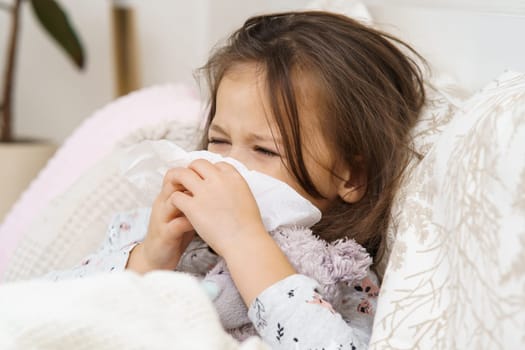 Little girl with closed eyes blow stuffy nose with handkerchief. Portrait of unhealthy child. Kid in sleepwear lie under blanket and have bed rest. Home treatment. Runny nose, snot, cold symptoms.