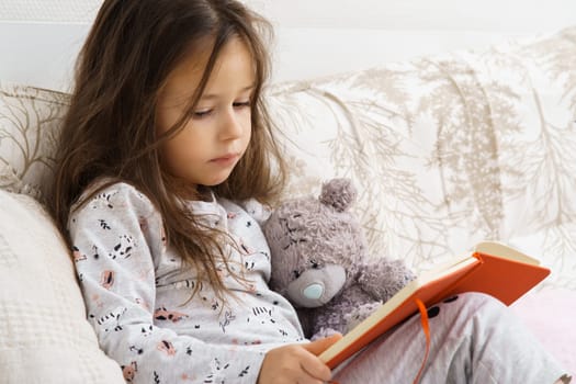 Unhappy little girl in sleepwear sitting on couch and reading book. Portrait of child, preschooler. Upset kid study at home. Hold storybook and teddy bear. Education, hobby, lifestyle.