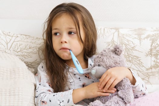 Sick little girl measure temperature with electronic thermometer in mouth and hug teddy bear. Portrait of unhealthy child. Unhappy kid in sleepwear have bed rest. Headache, fever heat, home treatment.