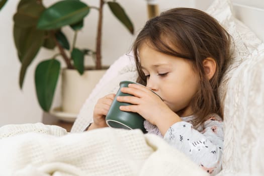 Nice little girl in sleepwear lying under blanket and drink warm beverage from cup. Portrait of unhealthy child, side view. Kid have bed rest. Home treatment, cold, disease. Taking medicament.
