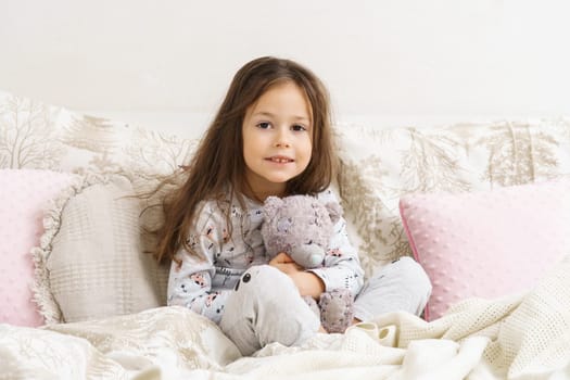 Little girl smiling with sleepy eyes and hugging favorite stuffed toy. Portrait of awaking child. Kid wake up at morning. Sleeping with stuffed toy, teddy bear. Resting on cozy bed, awakening.