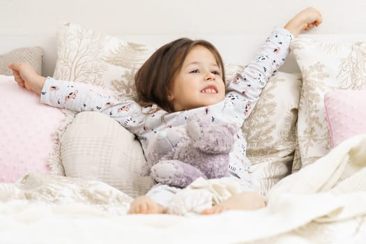 Happy little girl in sleepwear lying on bed and stretching with raised arms. Portrait of awaking child. Kid wake up at morning. Sleeping with stuffed toy, teddy bear. Resting in bedroom, awakening.
