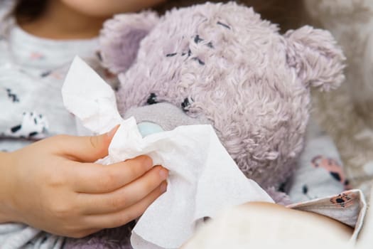 Hand of little girl in sleepwear blow nose of teddy bear with white handkerchief. Portrait closeup. Kid and toy have bed rest. Home patient treatment. Runny nose, cold symptoms, cure.