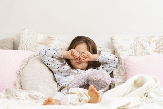 Adorable little girl in sleepwear lying on bed and rub eyes with fists. Portrait of awaking child. Kid wake up at morning. Sleeping with stuffed toy, teddy bear. Resting in bedroom, awakening.