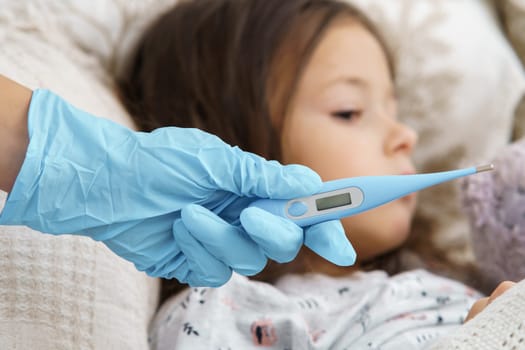 Doctor hand in blue medical glove hold electronic thermometer for measuring temperature of sick little girl. Selective focus, closeup view. Kid lie under blanket with fever heat and have bed rest.