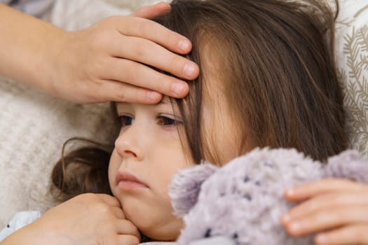 Hand of child touch hot forehead of sick little girl. Portrait closeup of unhealthy child. Kid with teddy bear lie with fever heat. Headache, high temperature. Treatment and bed rest.