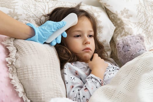 Doctor hand in medical glove measure temperature of sick little girl with electronic thermometer. Portrait of unhealthy child. Kid with teddy bear lie under blanket with fever heat. Cure and bed rest.