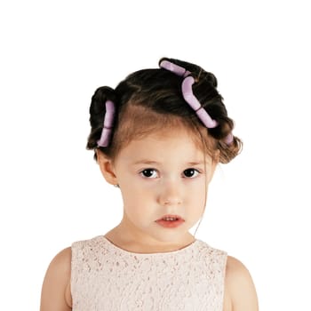 Discontent little girl with hair curlers. Portrait of child isolated on white. Kid in cute festive dress prepare hairstyle. Saint Valentine's Day concept, holiday outfit, unhappy emotions.