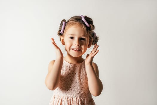 Joyful little girl with hair curlers. Portrait of smiling child on white. Kid in beautiful festive dress prepare hairstyle. Saint Valentine's Day concept, holiday outfit, happy emotions.