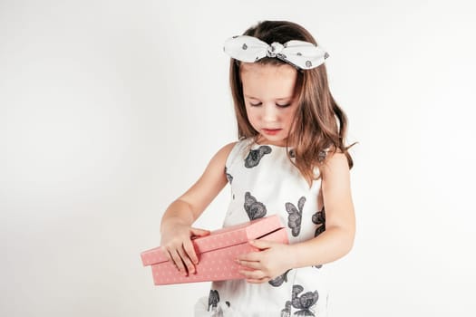 Adorable little girl open pink present box. Portrait of child on white. Pretty kid in festive dress and headband. Gift for holiday. Saint Valentine's Day concept, holiday of all lovers, celebration.