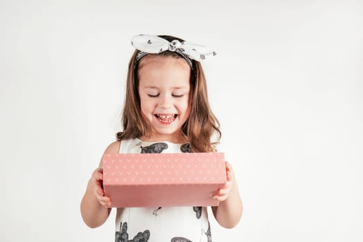 Joyful little girl hold pink present box looking down. Portrait of happy child on white. Pretty kid in festive dress and headband. Saint Valentine's Day concept, holiday of all lovers, celebration.