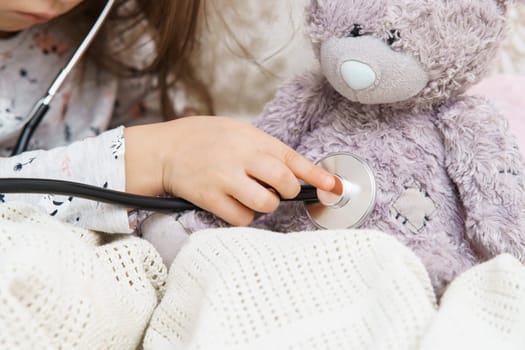 Hand of little girl listening to lung of teddy bear with stethoscope. Portrait of child closeup. Kid and toy sit under blanket and have bed rest. Home patient treatment. Caring doctor.