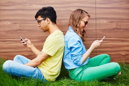Couple of teenagers sitting on green lawn with smartphones