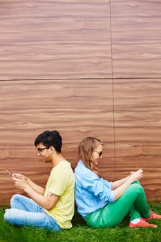 Couple of teenagers using modern gadgets while sitting on green lawn