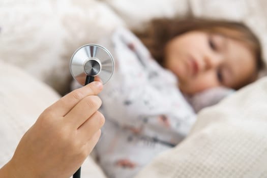 Human hand hold stethoscope for lung listening of little sick girl. Selective focus. Kid in sleepwear lie under blanket and have bed rest. Home patient treatment. Doctor, pediatrician.