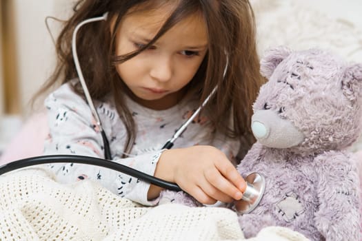 Serious little girl in sleepwear sitting under blanket and listening to lung of teddy bear with stethoscope. Portrait of child closeup. Kid and toy have bed rest. Home patient treatment.