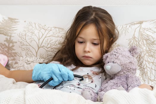Doctor hand in medical glove listening to lung of little sick girl with stethoscope. Portrait of unhealthy child. Kid in sleepwear lie under blanket with teddy bear and have bed rest. Home treatment.