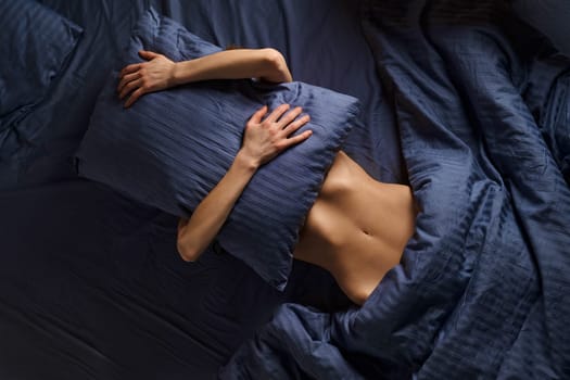 Concept of comfortable rest in bed, lazy weekend and introversion. Woman covers her face with pillow while lying in bed.