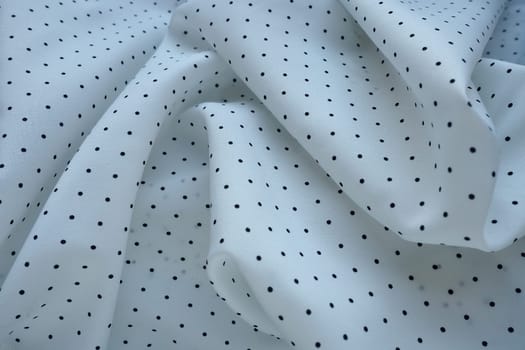 Texture of silk fabric. Black polka dots on a white background. Dress lightweight synthetic fabric in white with a repeating pattern. The textile is made up of waves and folds. Feminine classic style