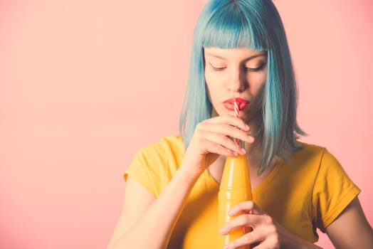 Closeup studio portrait of hipster young woman with red lipstick and blue dyed hair drinking orange juice from retro bottle with straw. Closeup of female model holding summer cocktail. Trendy pastel color background.