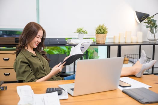Working on financial. Businesswoman sitting relaxing reclining with feet up on computer desk reading report document in paperwork clipboard, smiling woman holding papers document reading with laptop