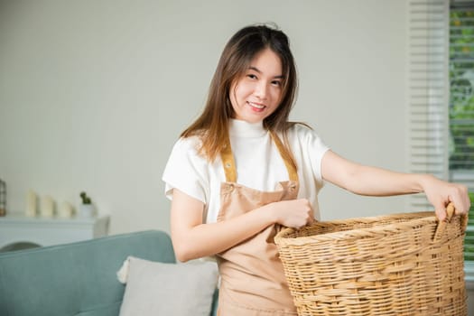 Beautiful housekeeper doing housework holds wooden basket of clean messy dirty clothes to washing at home, Asian housewife woman holding basket with heap of different clothes on sofa in living room