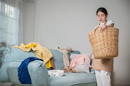 Beautiful housekeeper doing housework holds wooden basket of clean messy dirty clothes to washing at home, Asian housewife woman holding basket with heap of different clothes on sofa in living room