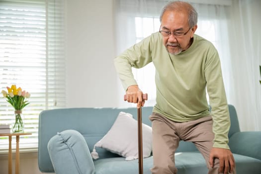 Asian Old man with eyeglasses typing to stand up from sofa with walking cane, Elderly suffering from knee pain ache holding handle of cane, senior disabled man holding walking stick at home